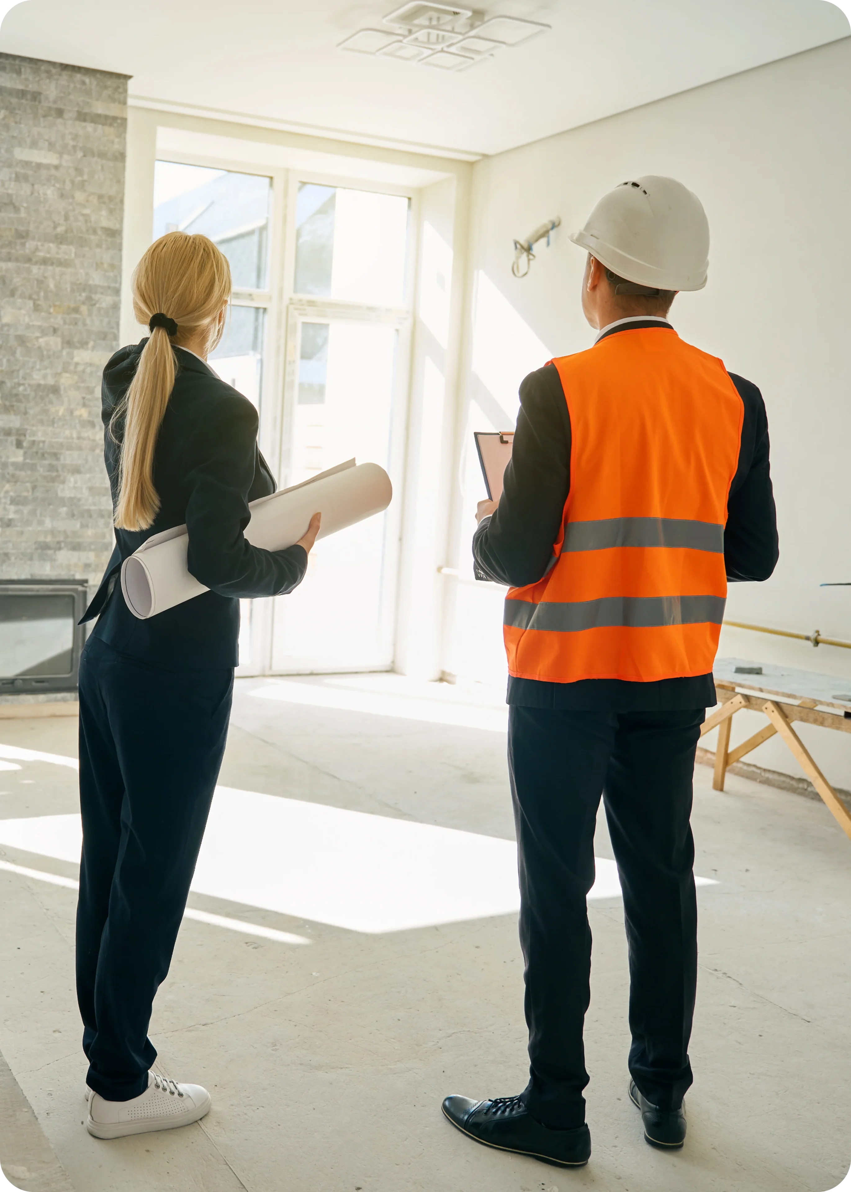 A woman holding blueprints and a man in a construction vest and helmet stand in a bright room with large windows, discussing plans.