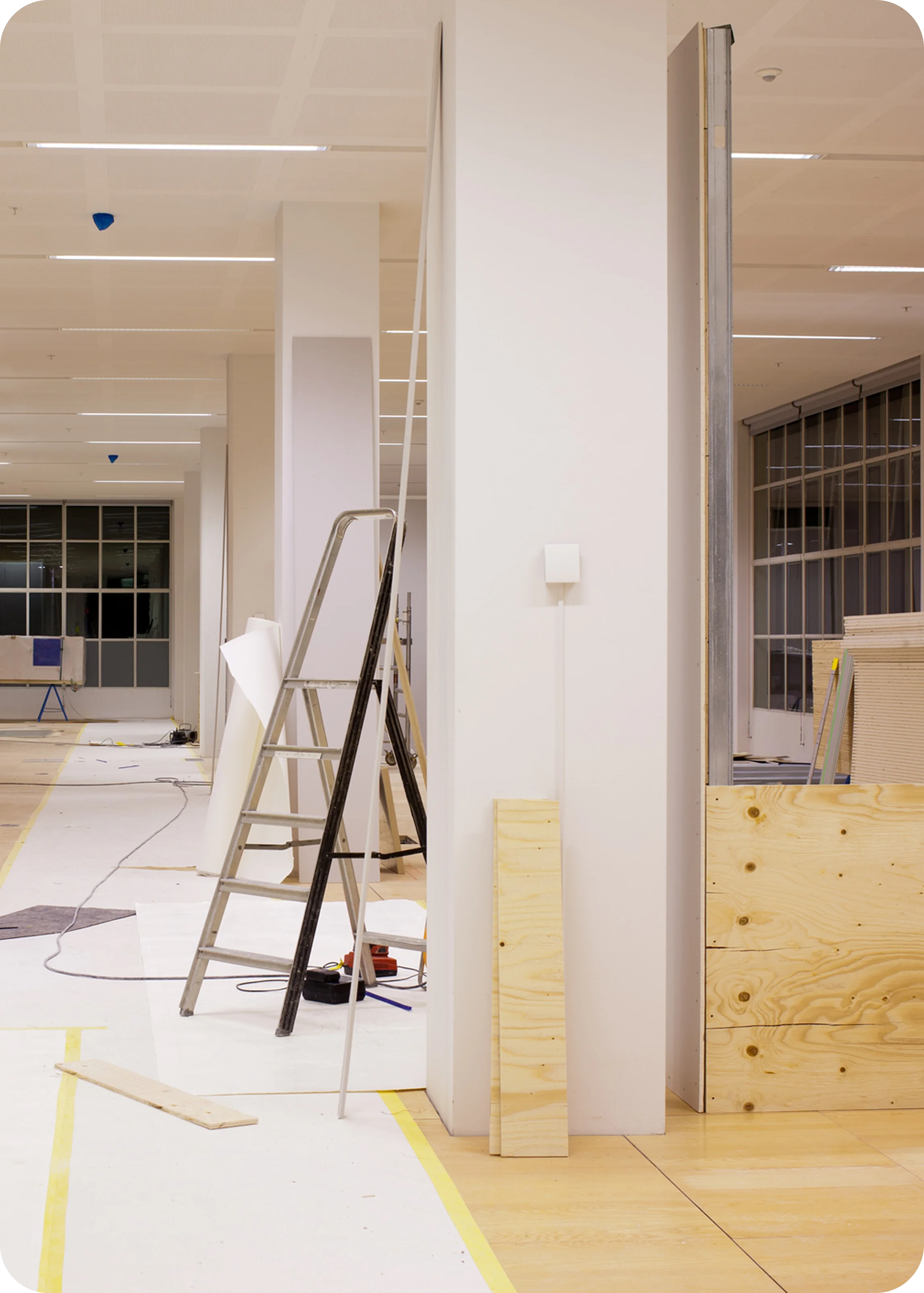 An indoor space under construction with a ladder, wooden planks, and construction materials scattered around. Fluorescent lights illuminate the area with a large window in the background.
